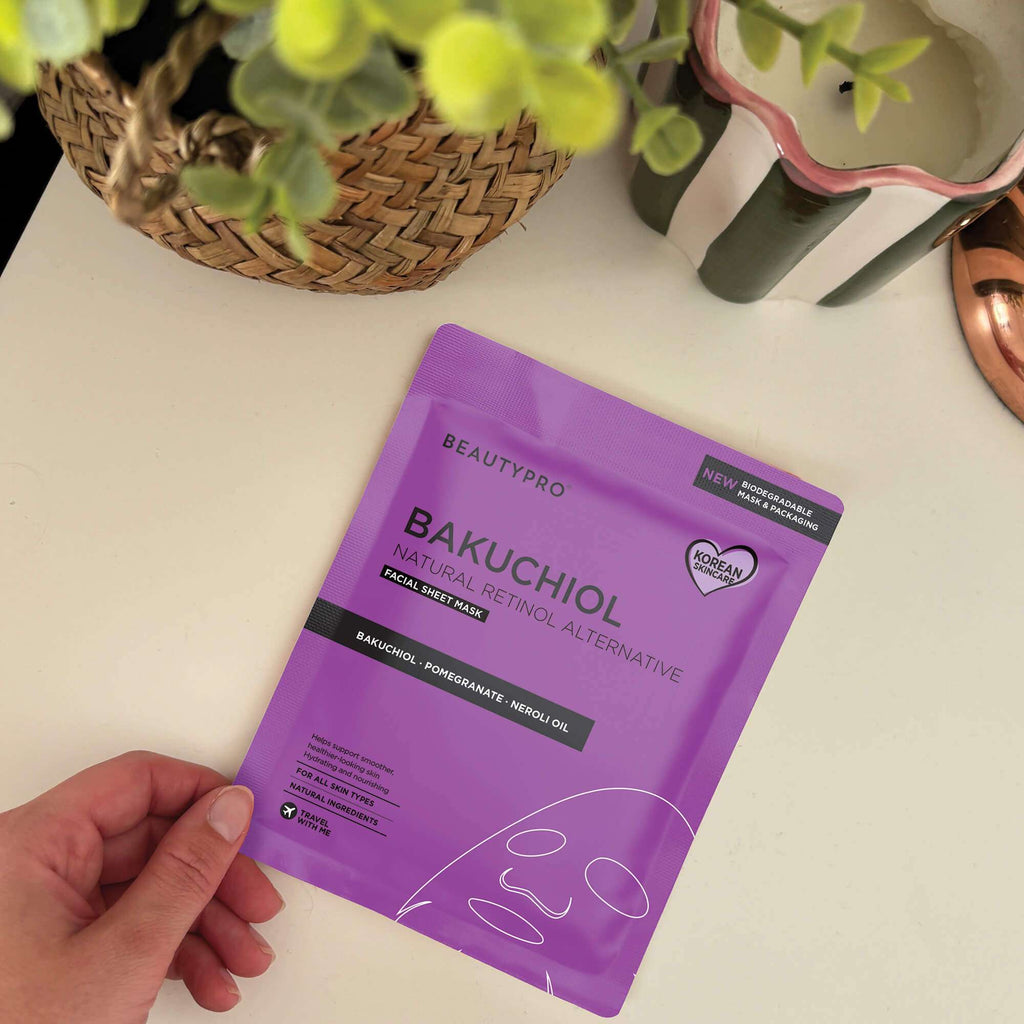 Hand holding a purple BeautyPro Bakuchiol facial sheet mask on a light surface with plants in the background.