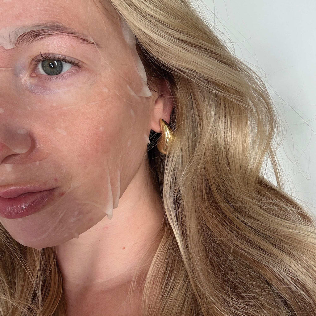 Close-up of a woman with blonde hair and a gold earring, wearing a white face mask.
