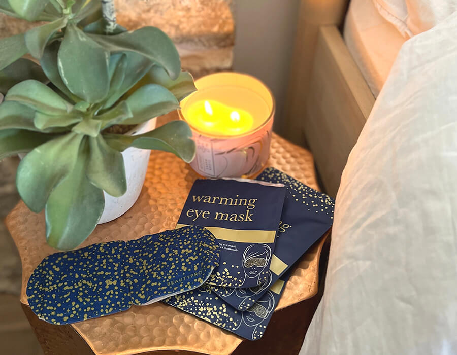 A warming eye mask and its packaging on a bedside table with a candle and a plant