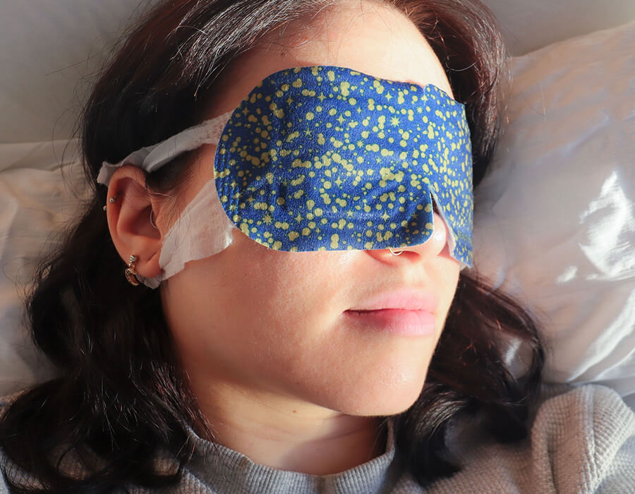 A warming eye mask being used by a woman asleep on a bed